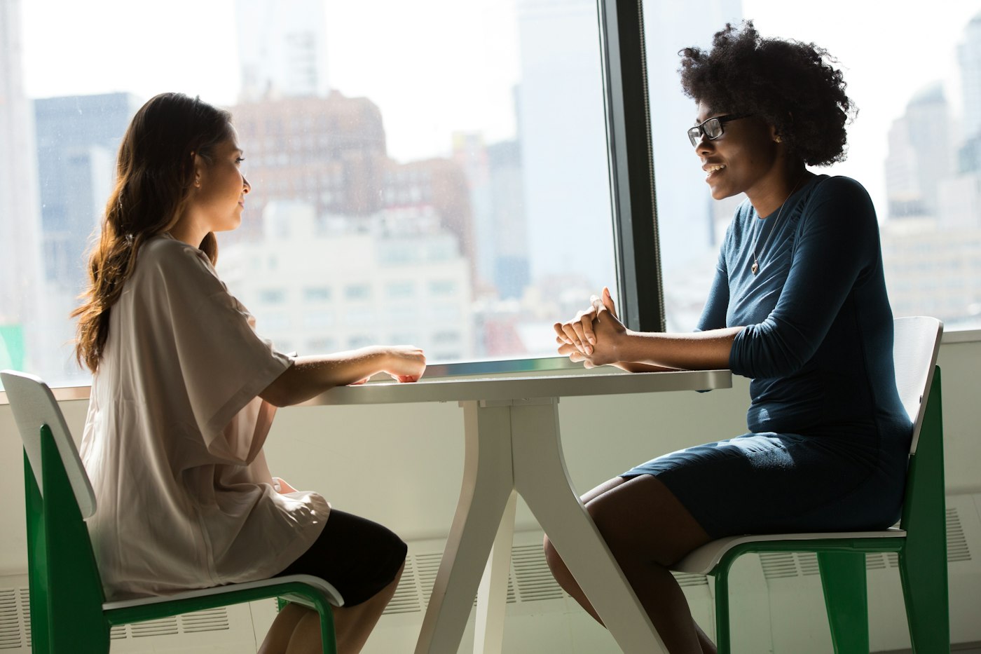 Two women talking across a small table in front of a large window